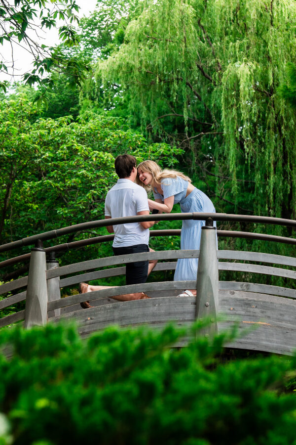 Joshua on one knee proposing to Hailey on a wooden footbridge surrounded by willow trees