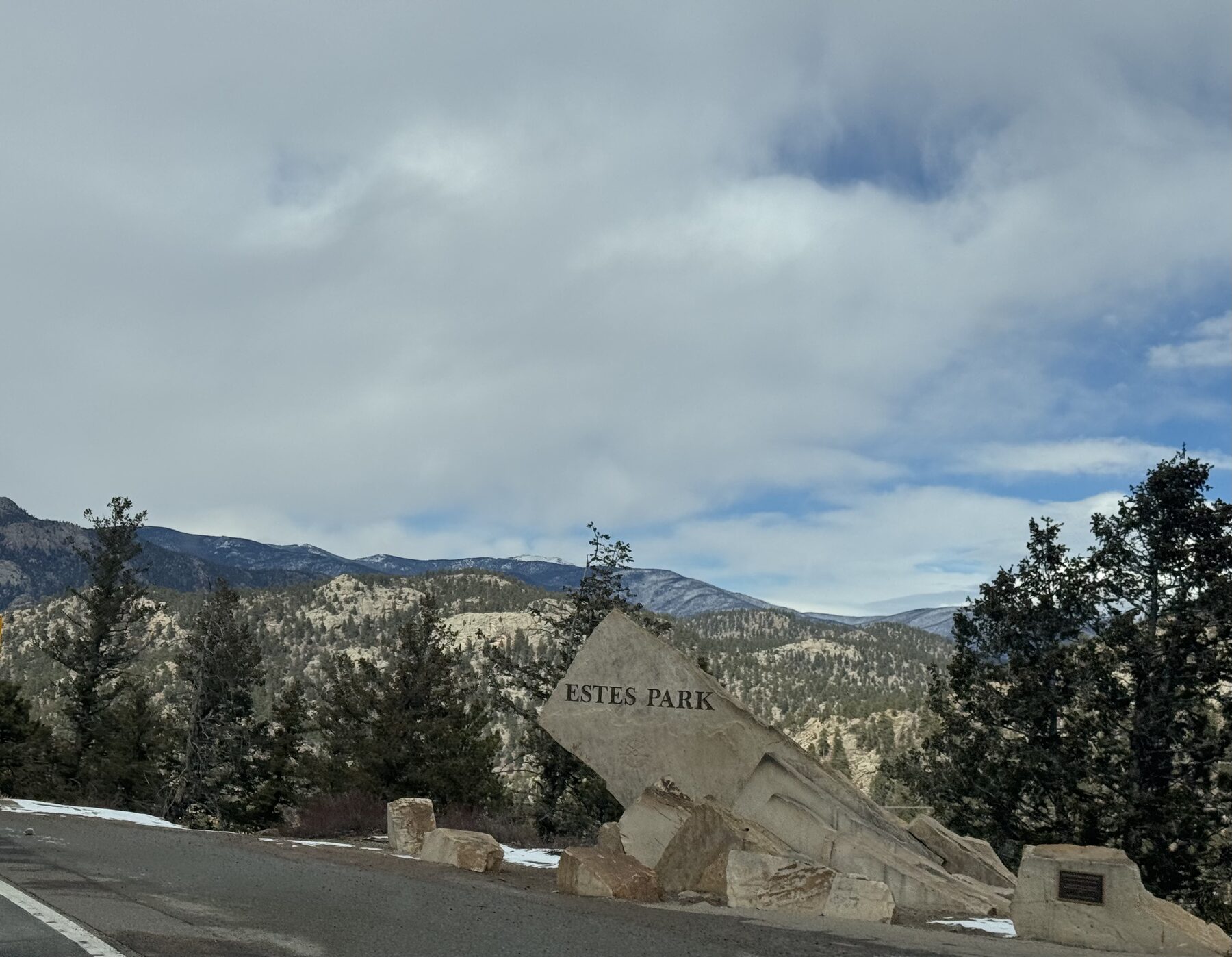 The carved stone Estes Park welcome monument beside the highway with snow-streaked foothills behind it