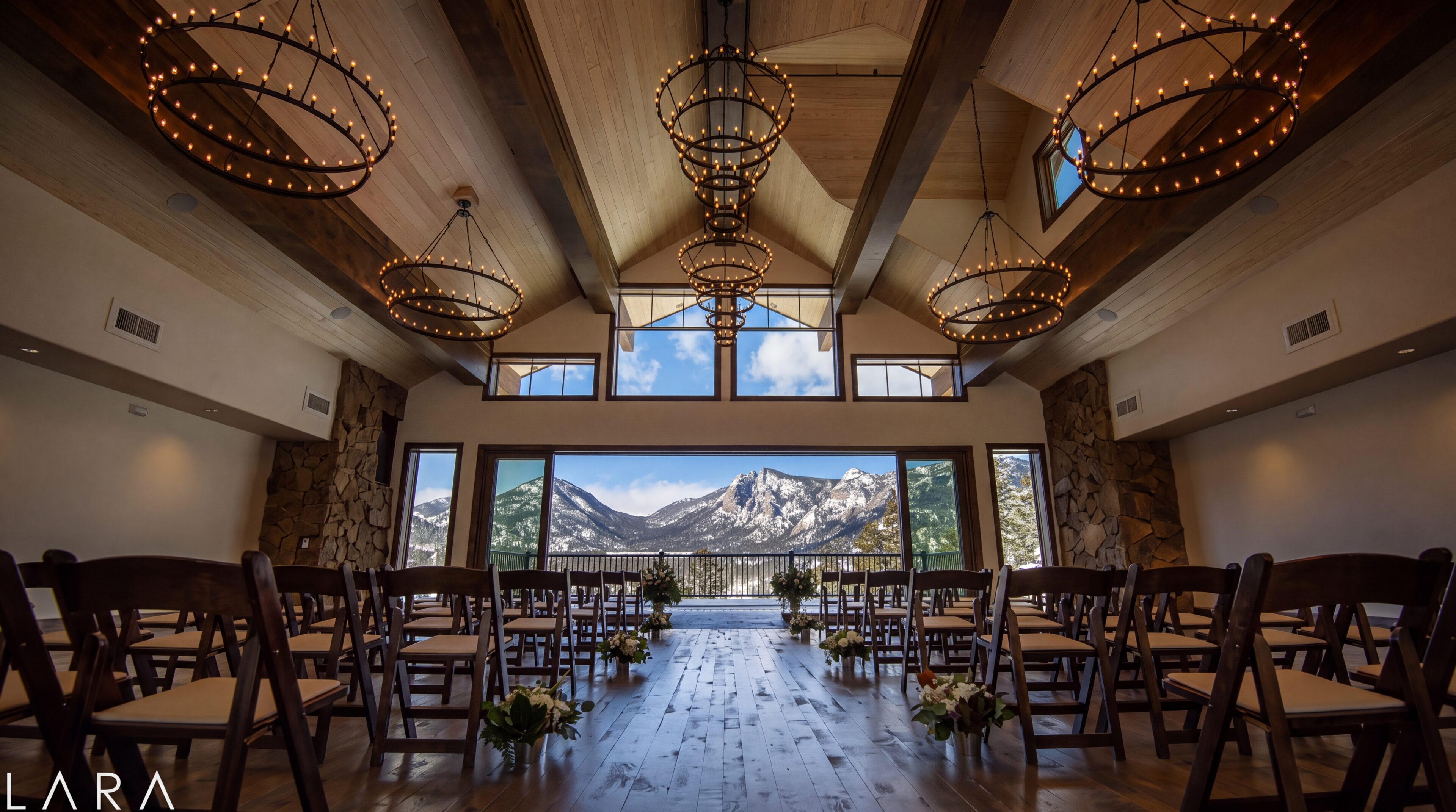 The ceremony hall at The Boulders: vaulted timber ceiling, ring chandeliers, wooden folding chairs, and a full glass wall opening onto snow-covered peaks